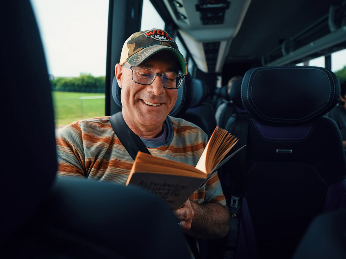 A gentleman in an orange and light blue striped shirt smiles and reads a book while riding the bus.