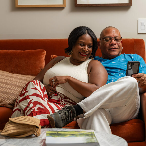 A sharply dressed couple sit on a vibrant burnt orange couch at Hotel Anna & Bel and smile at the cell phone.