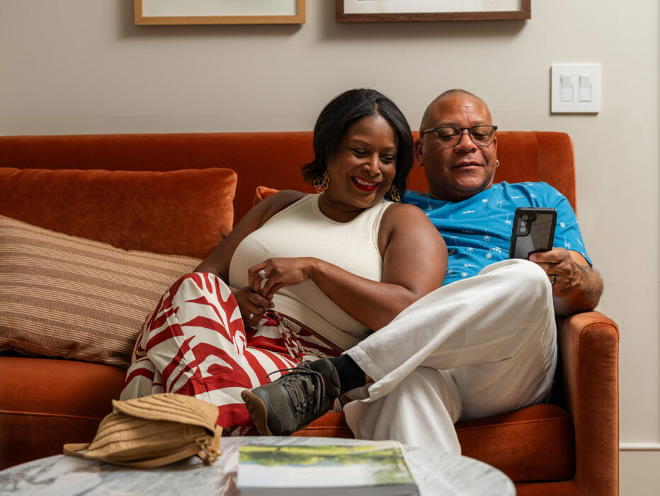 A sharply dressed couple sit on a vibrant burnt orange couch at Hotel Anna & Bel and smile at the cell phone.
