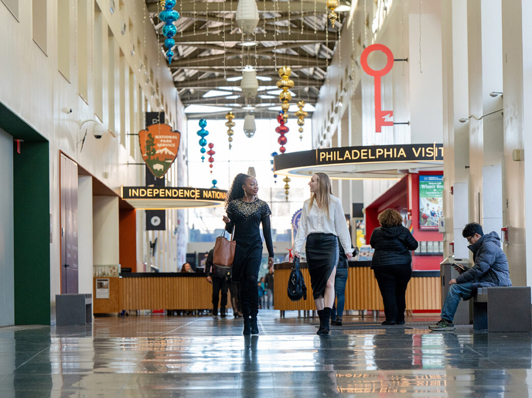 Two women walk through the decorated lobby of the Independence Visitor Center in Philadelphia, passing signs for the National Park Service and Philadelphia Visitor Center desks.