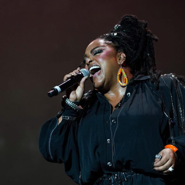 Jill Scott sings into a microphone while performing on stage. Her eyes are closed.