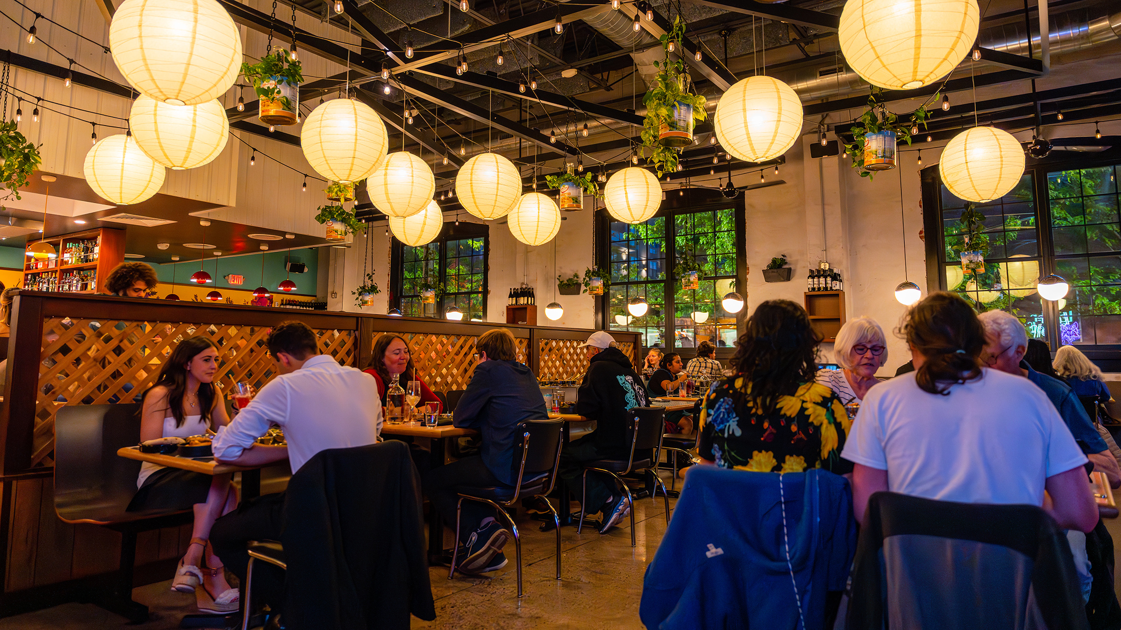Diners sit at closely spaced tables inside a warmly lit restaurant, with glowing paper lanterns, hanging plants and string lights overhead.