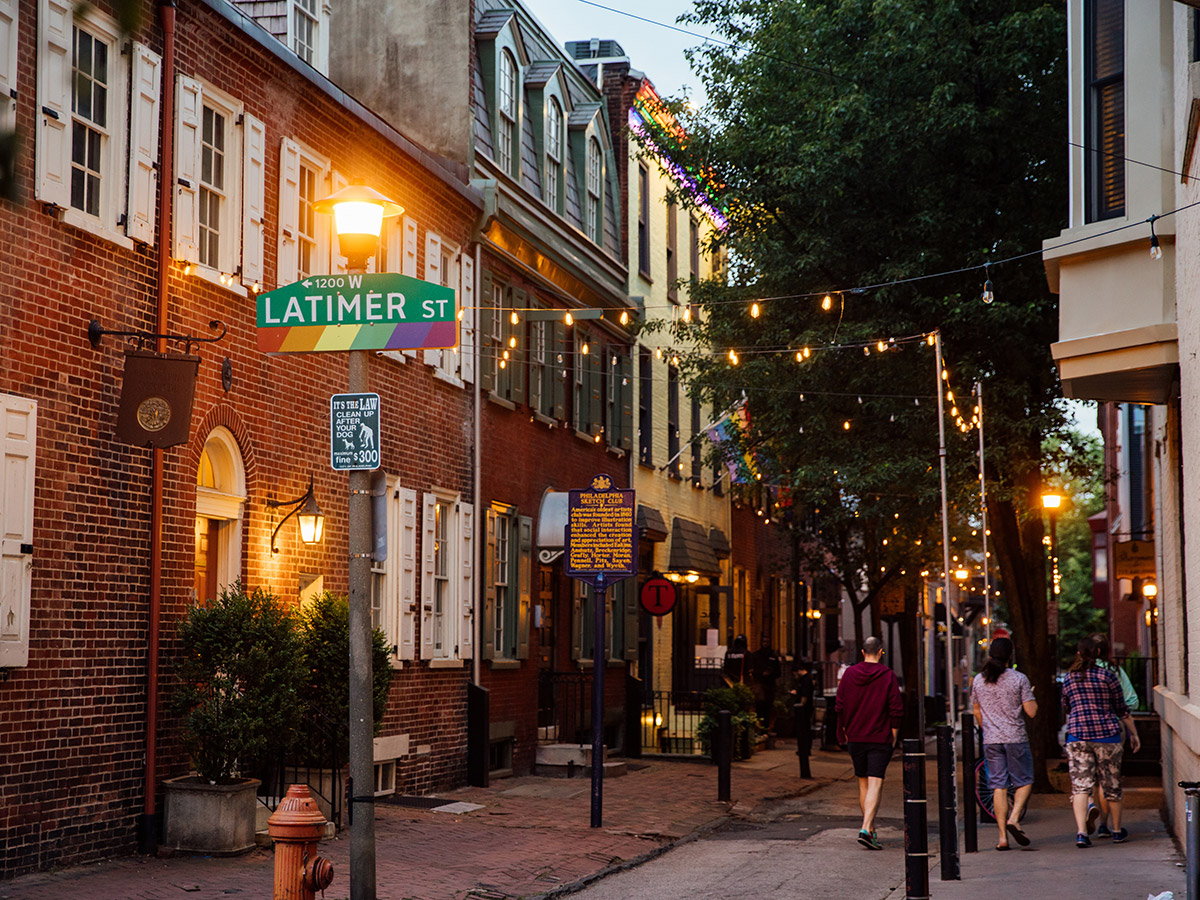 People dressed in shorts and t-shirts walk along Latimer Street at dusk under string lights, leafy trees and rainbow signage.
