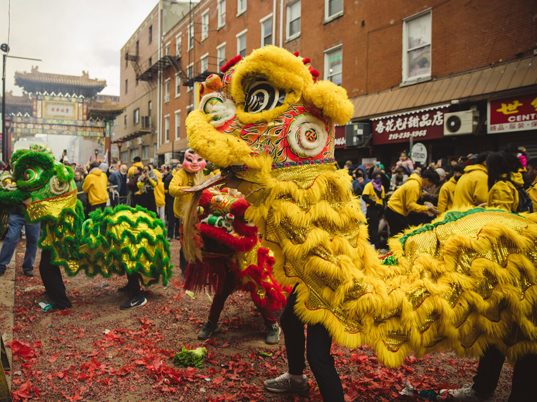 A vibrant lion dance performance fills a Chinatown street during Lunar New Year celebrations, with performers in bright yellow and green lion costumes surrounded by cheering crowds and red firecracker debris on the ground.