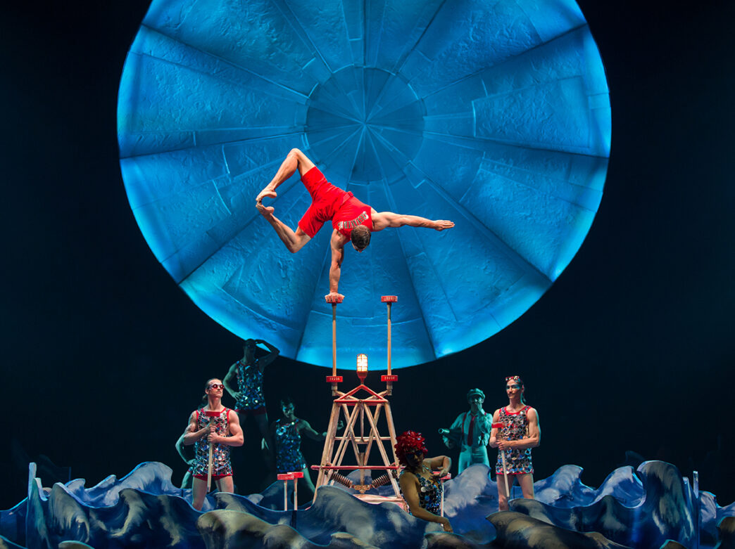 An acrobat balances upside down on a hand-held cane atop a platform, framed by a large glowing blue circle and surrounded by other performers on stage.