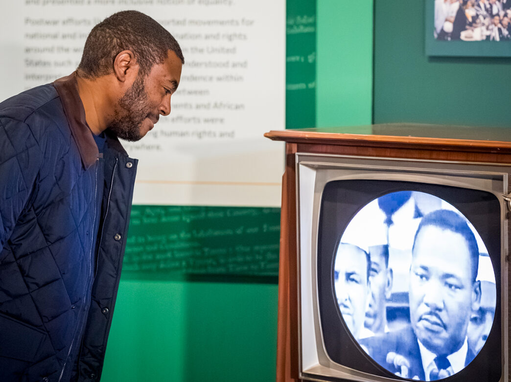 A man leans toward an old-fashioned television displaying black-and-white footage of Martin Luther King Jr., inside a museum style gallery with green walls.
