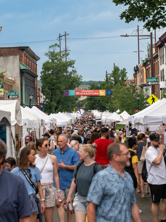 A large crowd shops at white vendor tents lining Main Street at the Manayunk Arts Festival.