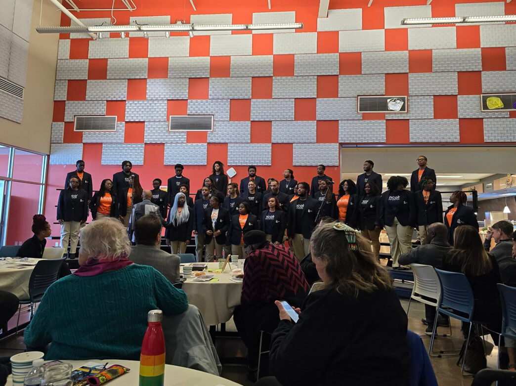 A choir stands in rows inside a bright community space with red-and-gray patterned wall behind them, performing for an audience seated at round tables.