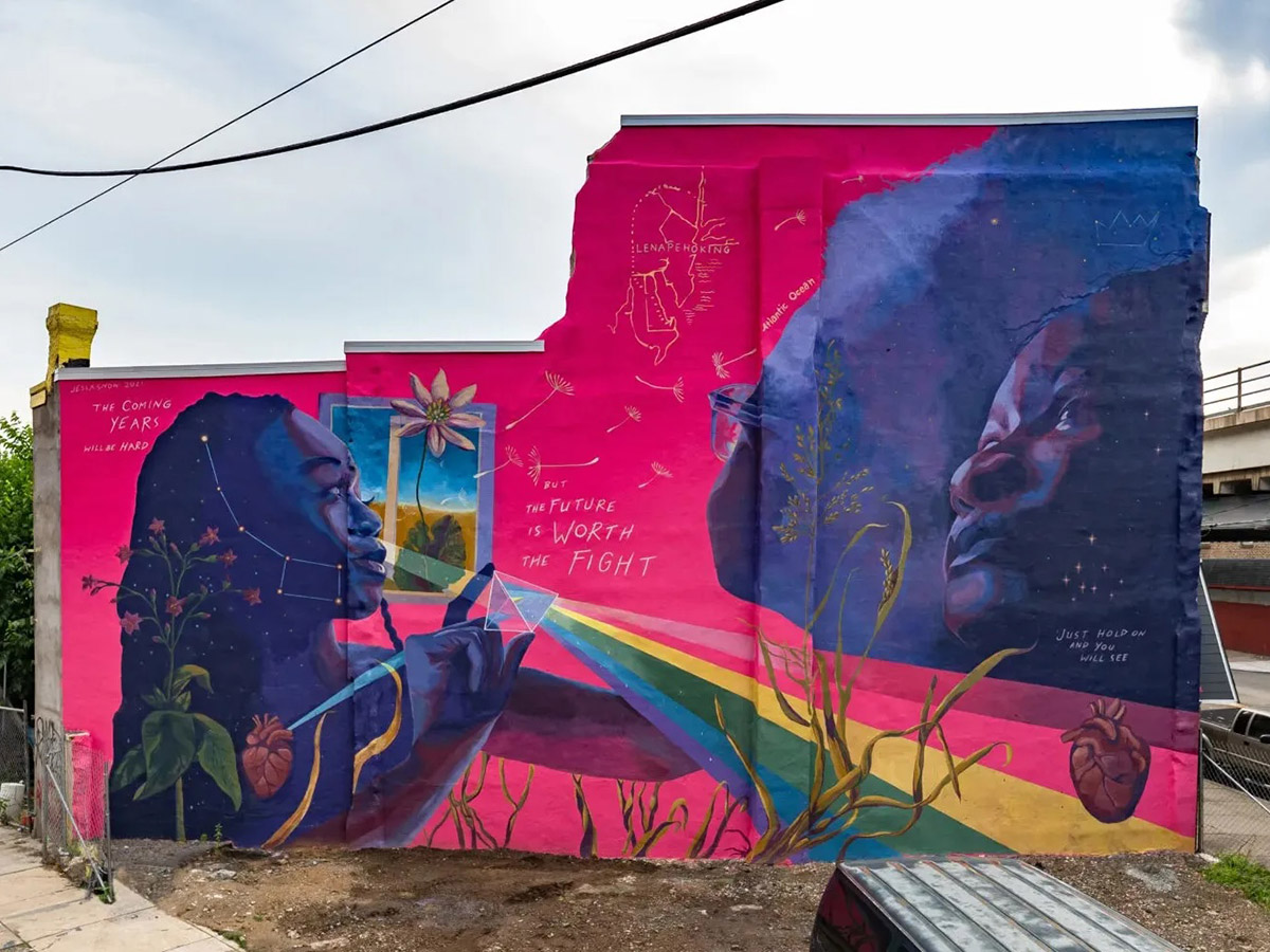 A vibrant mural of three young people painted in striking shades of blue and brown against a vivid pink background. Constellations of stars, anatomical hearts, flowers, rainbows and the phrase "The coming years will be hard, but the future is worth the fight. Just hold on, and you will see," are also seen in the mural.