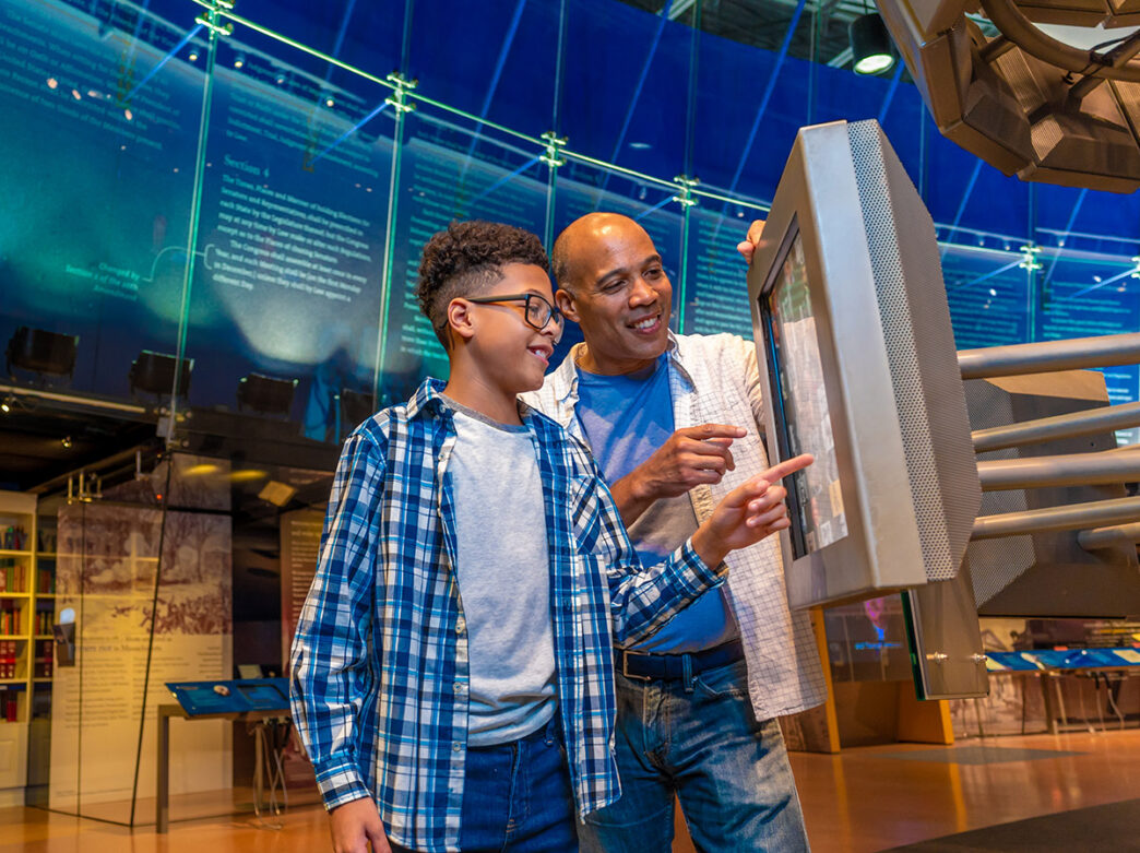 A smiling father dressed in a white button-up shirt and his son dressed in a plaid blue shirt explore an interactive touchscreen display at the National Constitution Center.