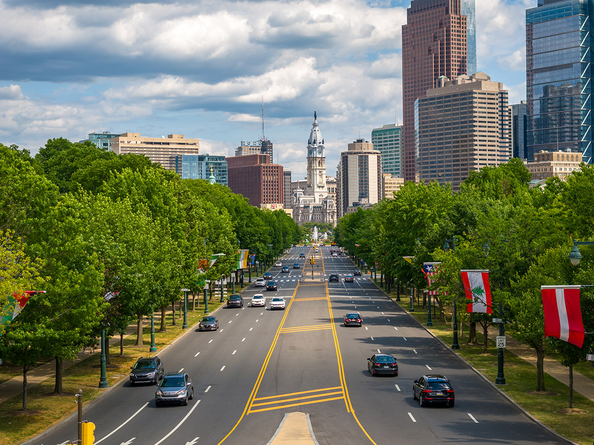 A photo of the Benjamin Franklin Parkway. Lush, green trees line the street, cars of a variety of models drive up and down the street, and City Hall can be seen in the background.