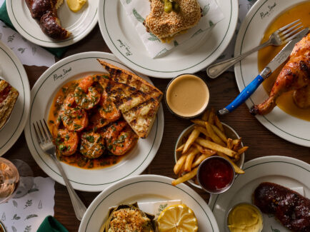 An overhead view shows an spread of a restaurant dishes, including roasted chicken, sandwiches, saucy shrimp, French fries and more.