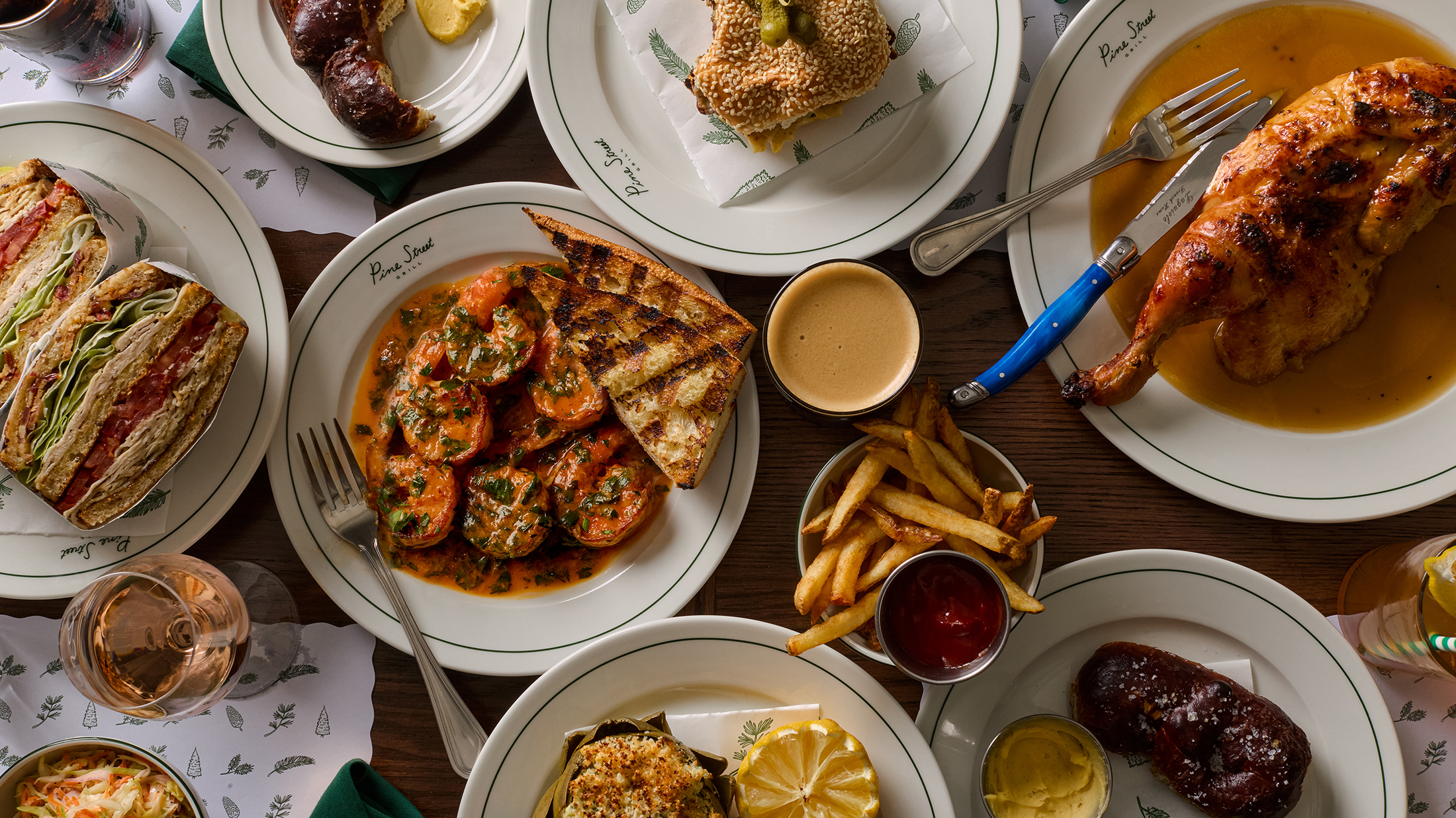 An overhead view shows an spread of a restaurant dishes, including roasted chicken, sandwiches, saucy shrimp, French fries and more.