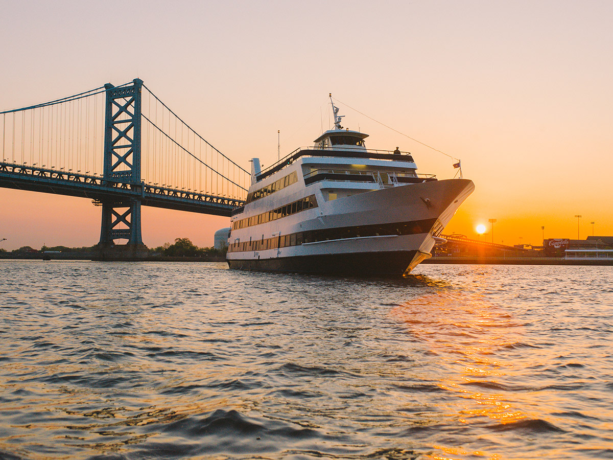 The Spirit of Philadelphia, a large white and black cruise ship, sales down the Delaware River as the sun sets. The Benjamin Franklin Bridge can be seen in the background.
