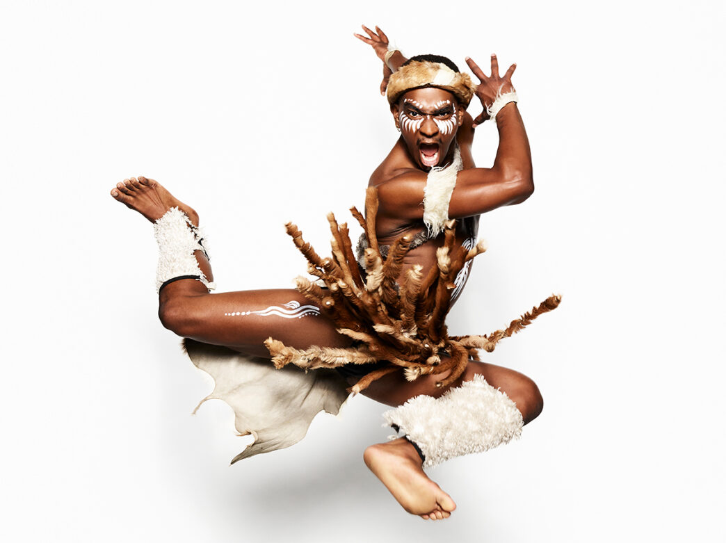 A Step Afrika! dancer leaps midair against a white background while wearing a traditional costume with white face paint and fur accents.