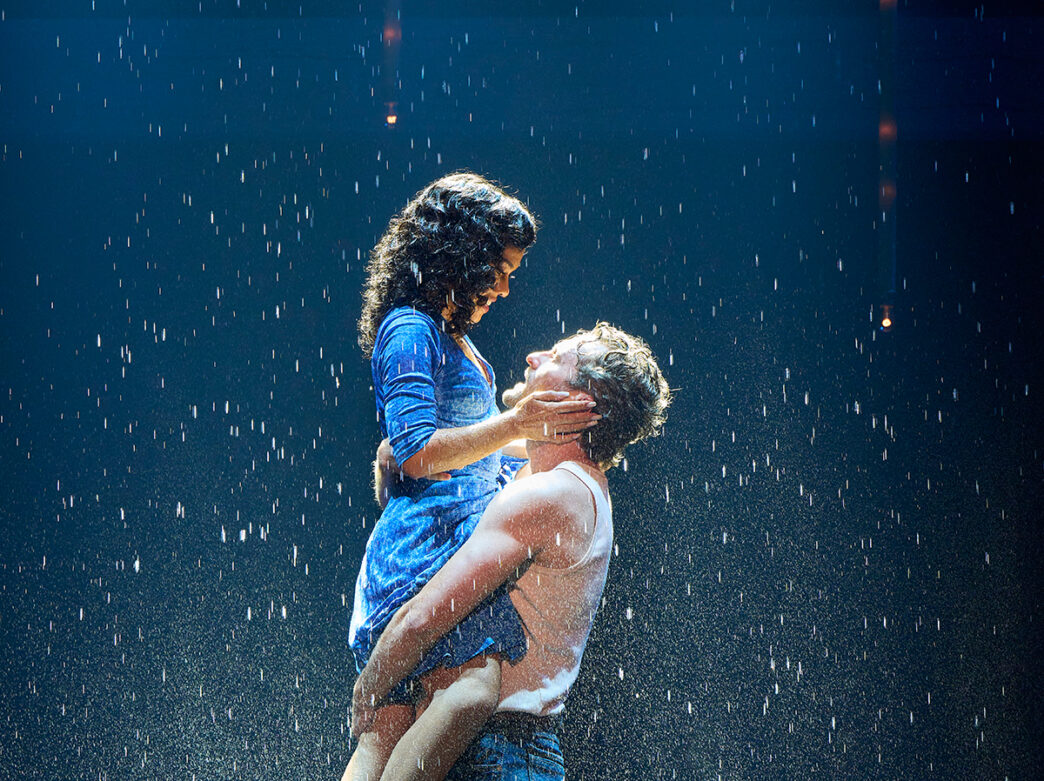 A man wearing a white tank top lifts a woman into the air onstage as rain falls around them, the two gazing at each other under dramatic blue lighting.