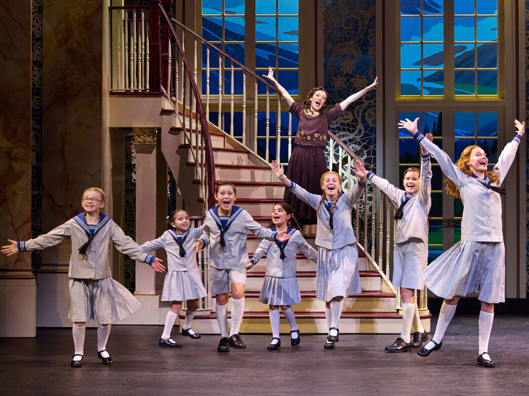 A group of children in matching light blue outfits sing and dance onstage with arms raised, while an adult performer stands singing behind them on a staircase.