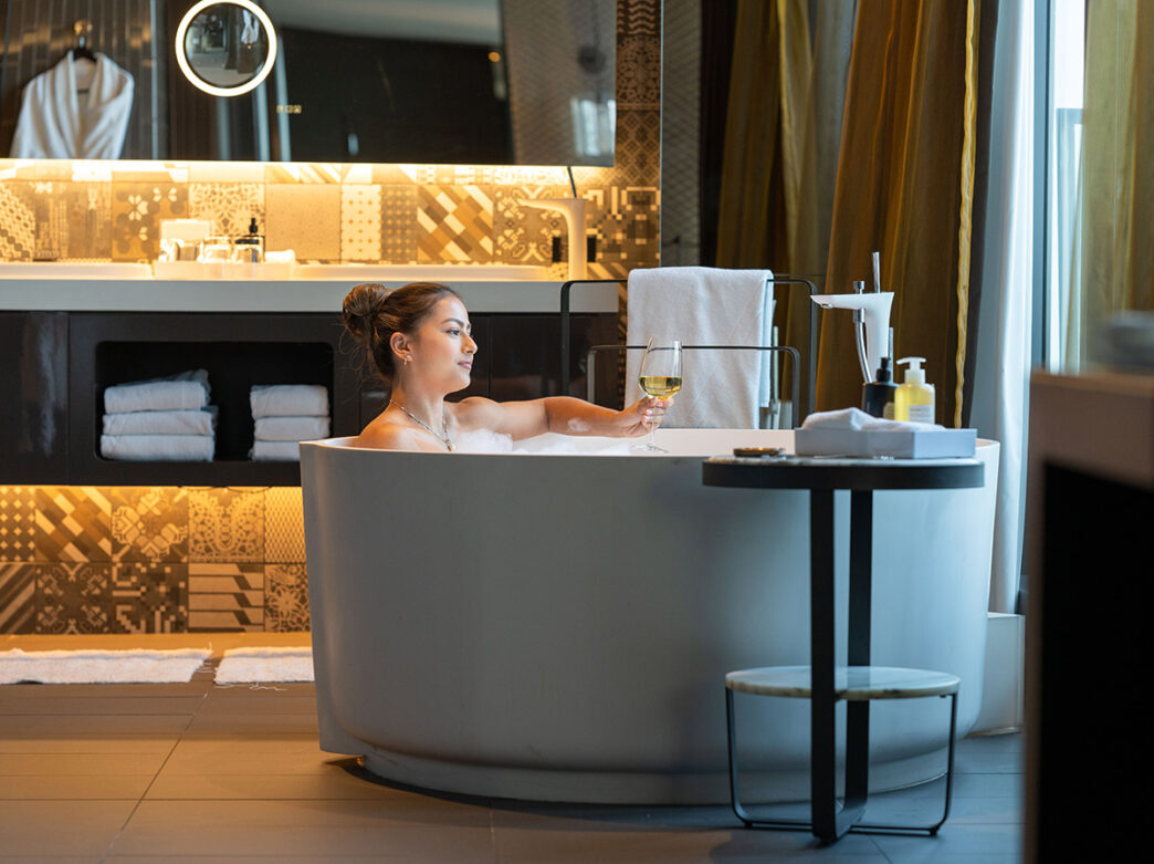 A woman holds a glass of wine as she soaks in a modern and minimalistic in-room bathtub at The W Philadelphia.