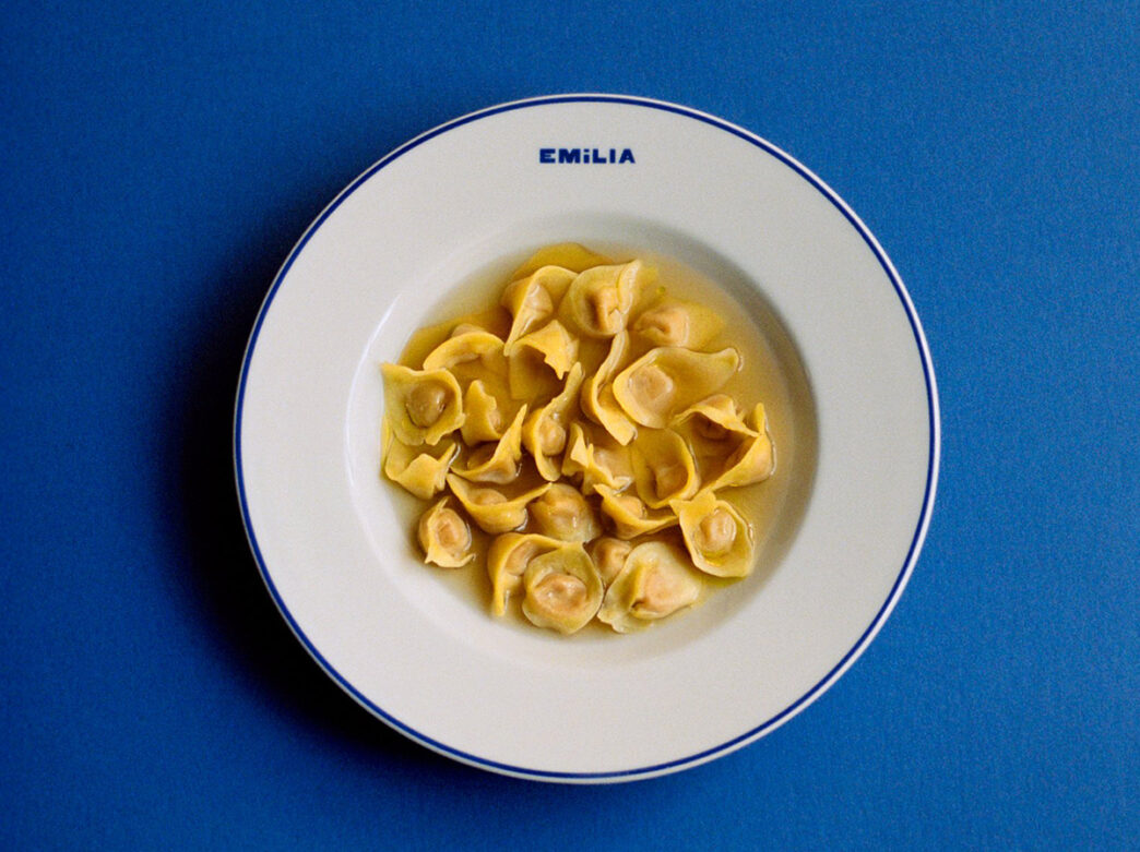 A white bowl holds tortellini en brodo, with delicate stuffed pasta floating in golden broth against a deep blue background.