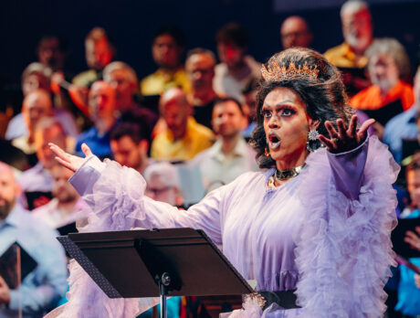 Drag queen Cookie DiOrio zingt jubelend voor een koor van mannen. De koningin draagt een kroon en een paarse jurk met franjes.