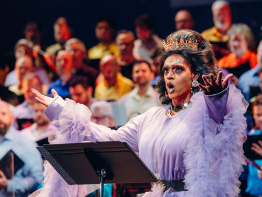 Drag queen Cookie DiOrio sings jubilantly in front of a chorus of men. The queen wears a crown and a frilled purple dress