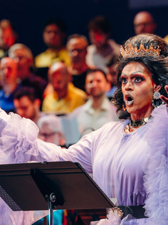 Drag queen Cookie DiOrio sings jubilantly in front of a chorus of men. The queen wears a crown and a frilled purple dress