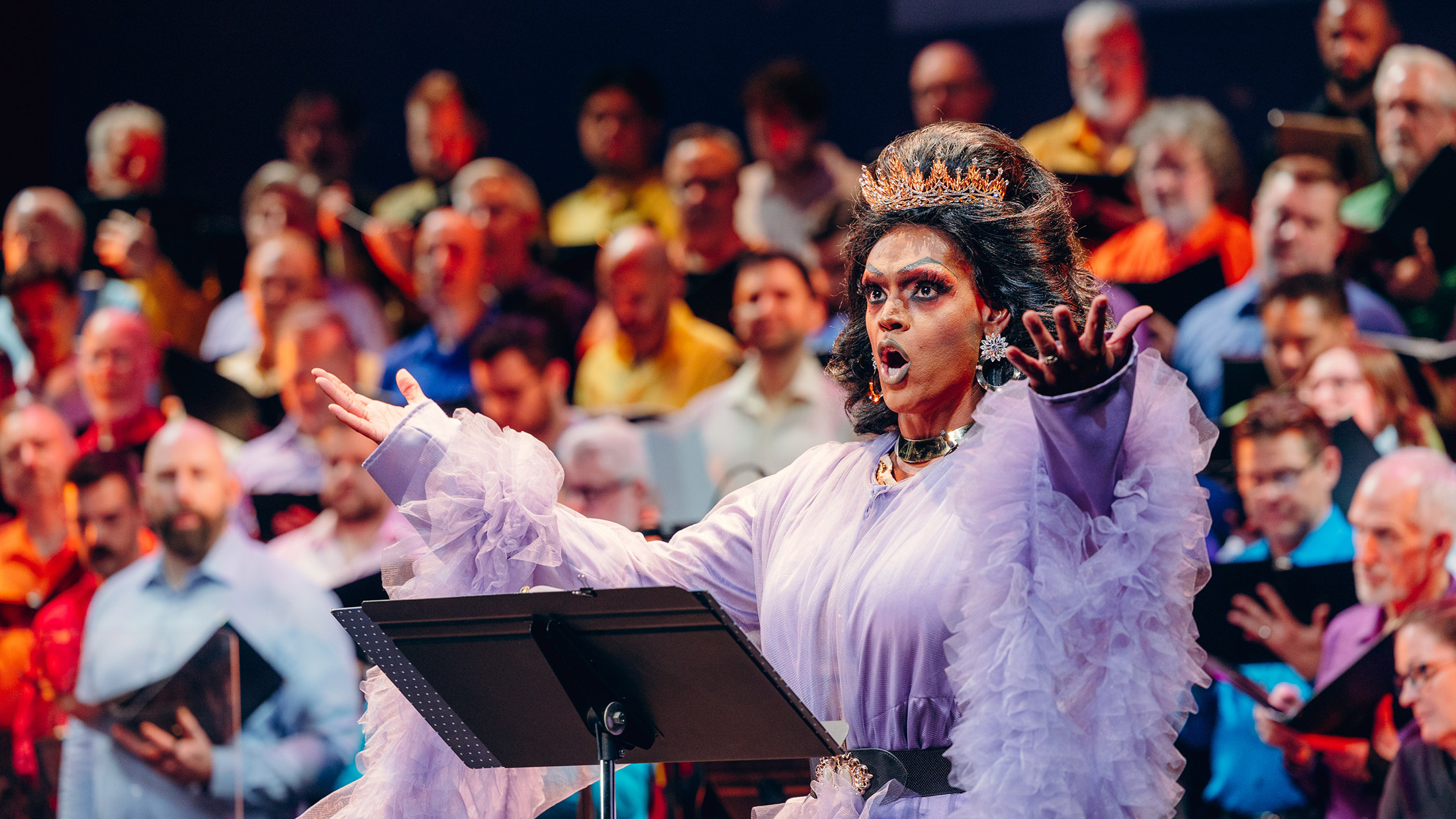 Drag queen Cookie DiOrio sings jubilantly in front of a chorus of men. The queen wears a crown and a frilled purple dress
