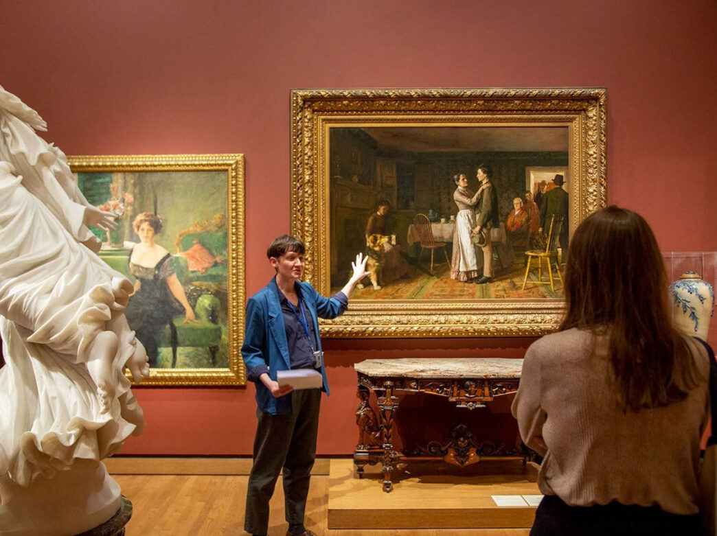 Een museumgids wijst naar een oud schilderij van mensen die in een historisch huis staan. Een bezoeker observeert de gids en de galerie van een afstandje