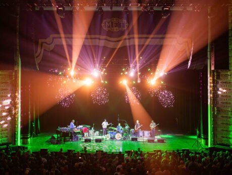 A band performs on stage at an indoor theater as colorful light beams illuminate the musicians and a large crowd fills the floor in front of the stage.
