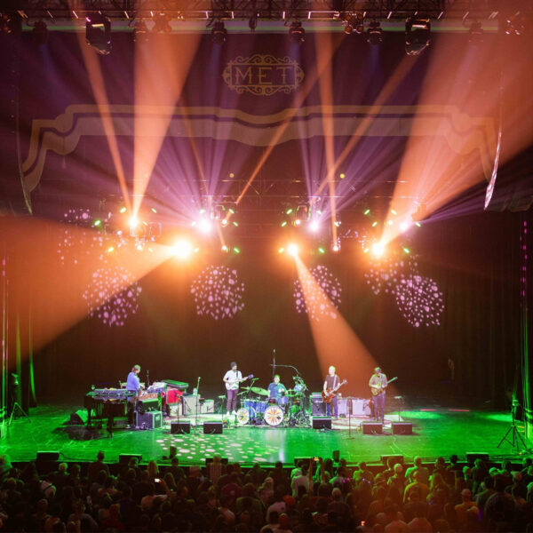 A band performs on stage at an indoor theater as colorful light beams illuminate the musicians and a large crowd fills the floor in front of the stage.