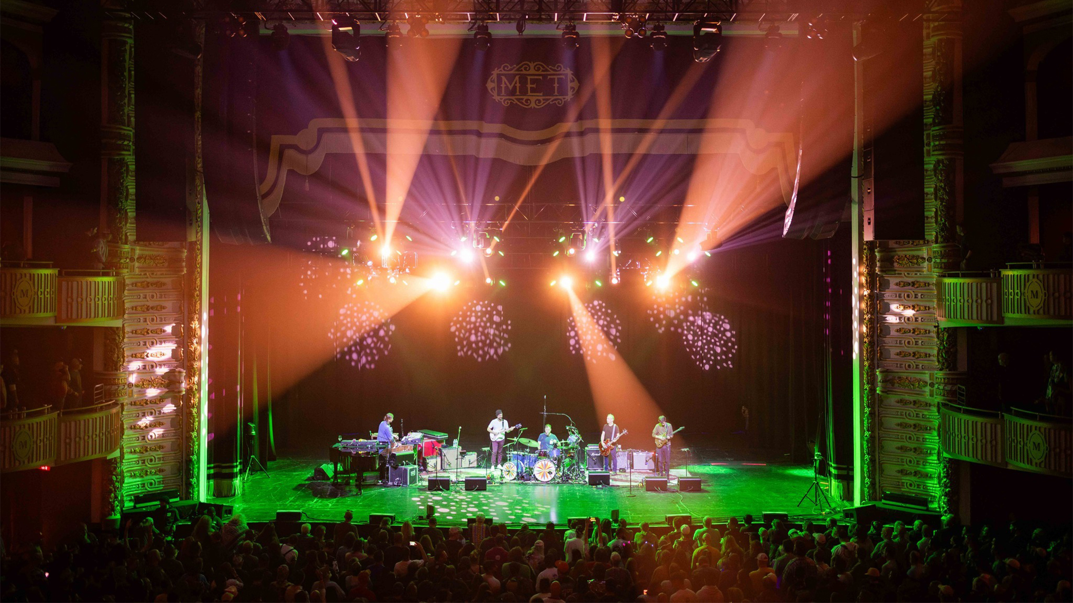 A band performs on stage at an indoor theater as colorful light beams illuminate the musicians and a large crowd fills the floor in front of the stage.
