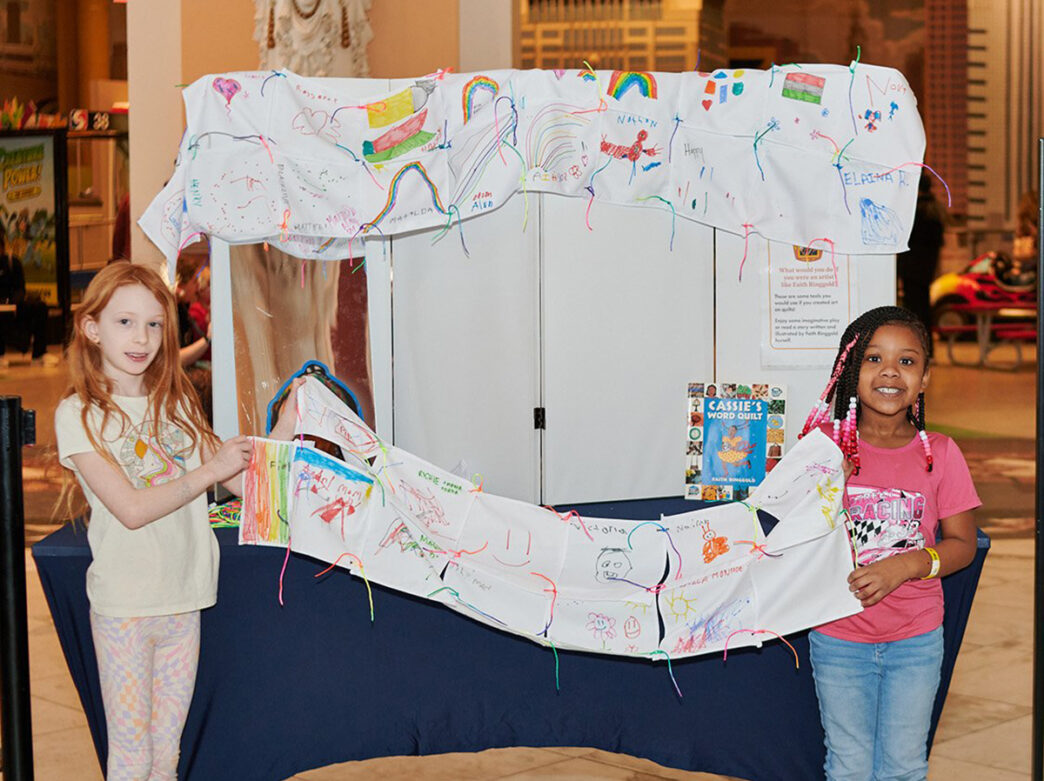 Two children stand on either side of a long fabric banner covered in colorful drawings, proudly displaying their artwork at an indoor museum activity table.