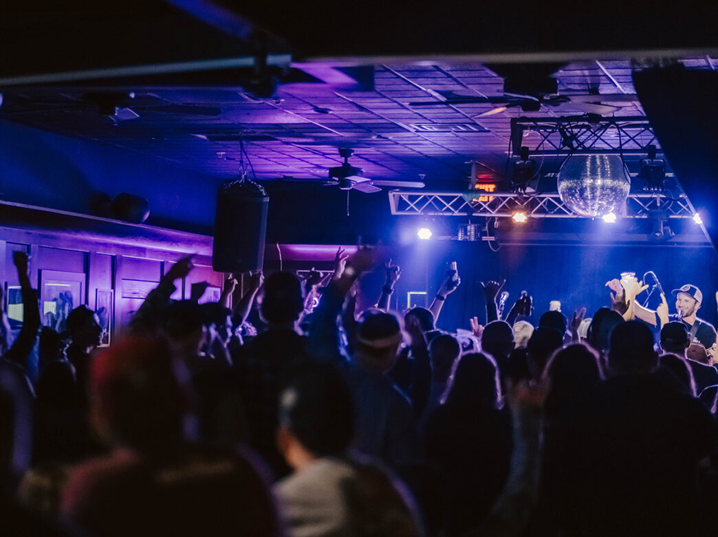 Los asistentes levantan las manos al aire frente a un escenario bañado por luces moradas y azules, con una bola de discoteca colgando sobre sus cabezas durante un concierto íntimo.
