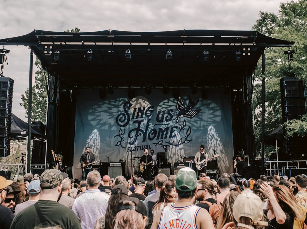 A band performs on an outdoor festival stage with "Sing Us Home" printed on the backdrop as a large crowd watches from the lawn.
