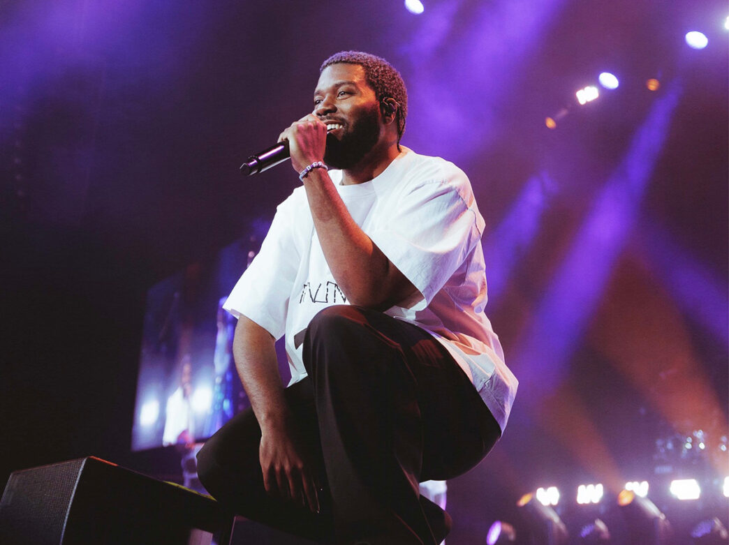 A singer kneels on stage holding a microphone up to their mouth while performing. Purple and blue concerts lights illuminate the stage.