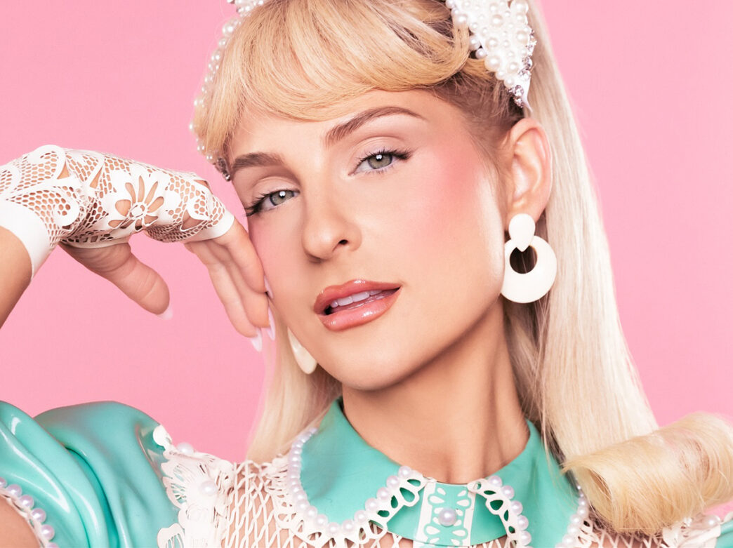 A pop singer poses against a pink background wearing a pastel teal dress, pearl accessories and lace gloves, rest her head on one hand.