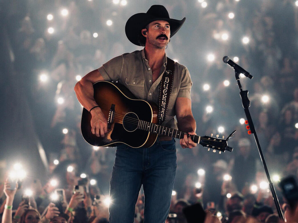 A musician wearing a cowboy hat plays an acoustic guitar onstage as a large crowd holds up glowing phone lights in the background.