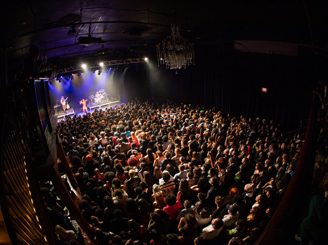 Una multitud abarrotada llena una oscura sala de conciertos, vista desde arriba, mientras los músicos tocan en un escenario brillantemente iluminado bajo una lámpara de araña.
