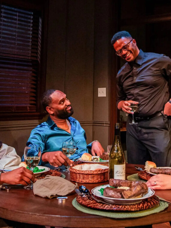 Four actors sit and stand around a dining table set with food and wine, smiling and laughing together in an intimate stage scene.