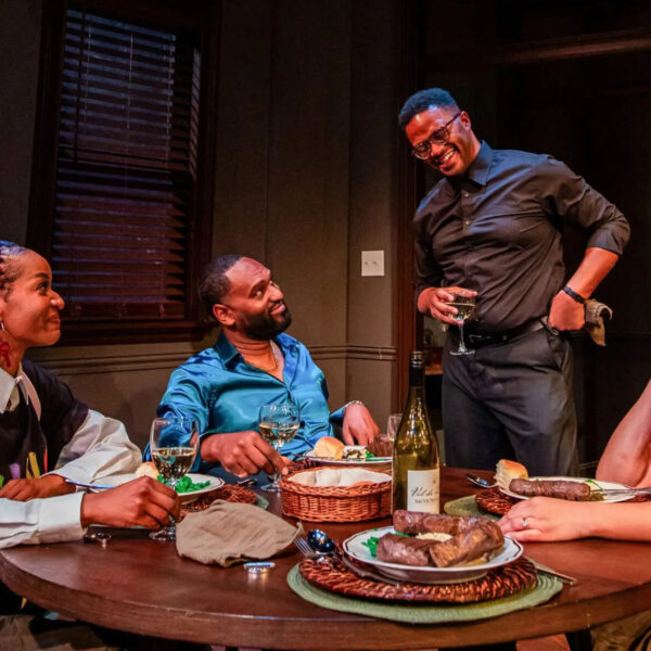 Quatro atores sentam-se e ficam em pé ao redor de uma mesa de jantar com comida e vinho, sorrindo e rindo juntos em uma cena íntima no palco.