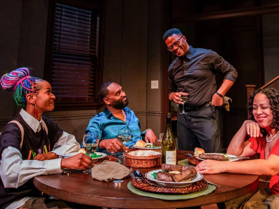 Four actors sit and stand around a dining table set with food and wine, smiling and laughing together in an intimate stage scene.