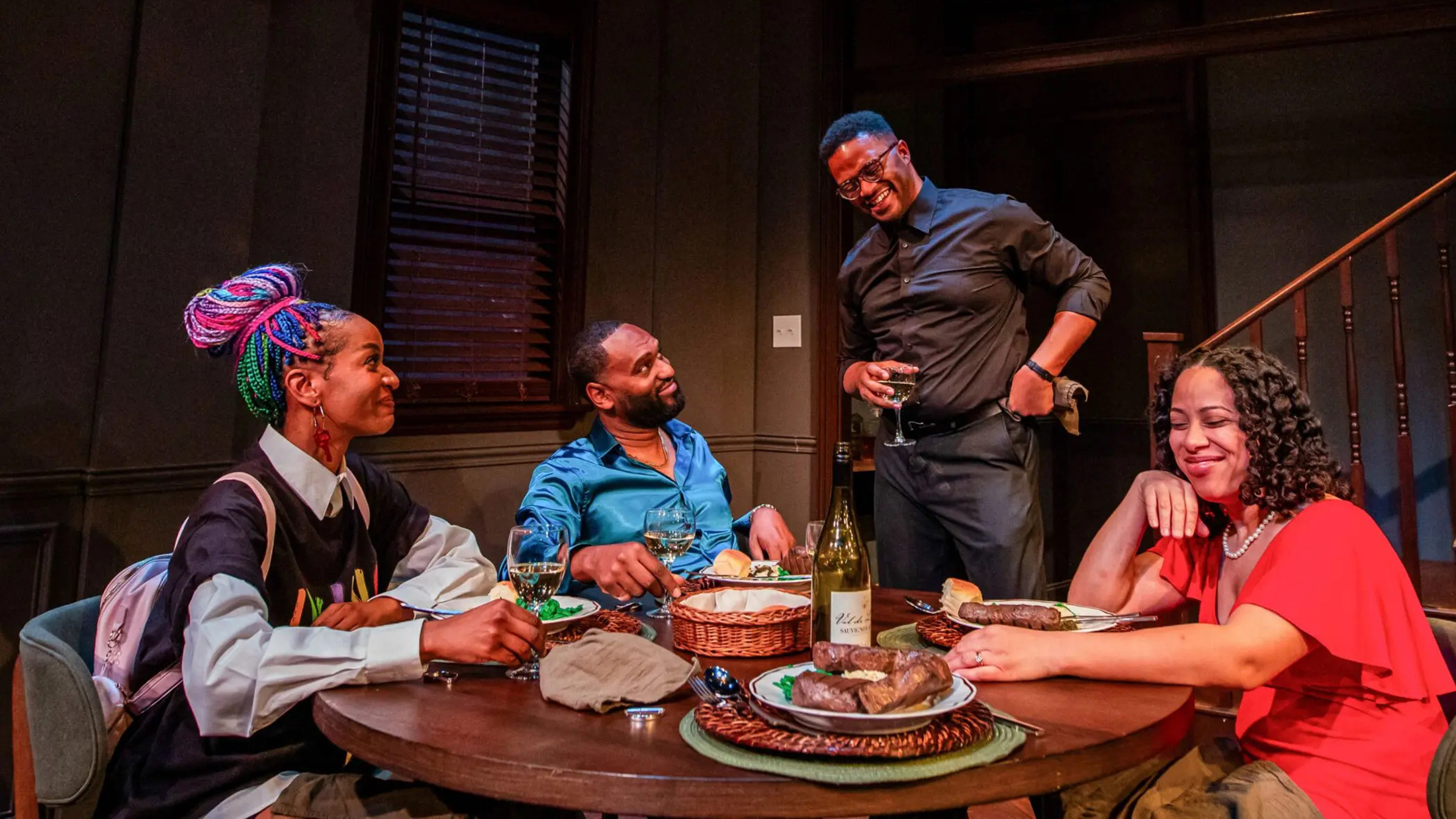 Four actors sit and stand around a dining table set with food and wine, smiling and laughing together in an intimate stage scene.