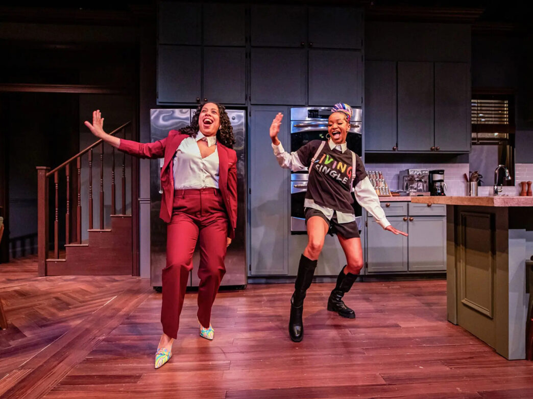 Two actors dance joyfully in a kitchen set on stage, one in a red suit and the other in a casual outfit, both smiling with arms raised mid-movement.