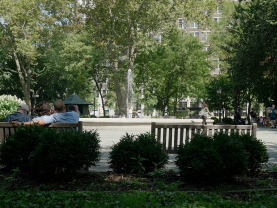 Three people sit on a park bench facing a fountain in Philadelphia’s leafy Rittenhouse Square, enjoying a sunny day while other visitors walk around the fountain.