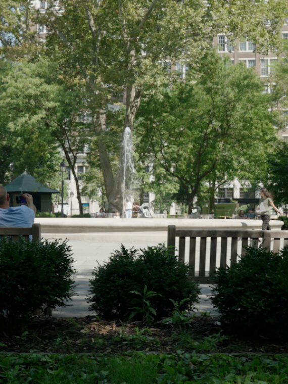 Three people sit on a park bench facing a fountain in Philadelphia’s leafy Rittenhouse Square, enjoying a sunny day while other visitors walk around the fountain.