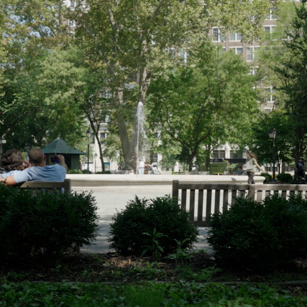 Three people sit on a park bench facing a fountain in Philadelphia’s leafy Rittenhouse Square, enjoying a sunny day while other visitors walk around the fountain.
