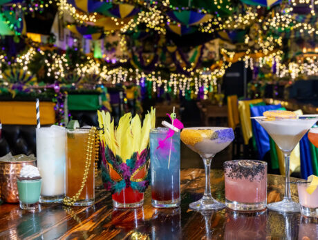 A lineup of colorful Mardi Gras-themed cocktails sit on a wooden table inside a festively decorated restaurant, with beads, feathers and purple, green and gold decor.