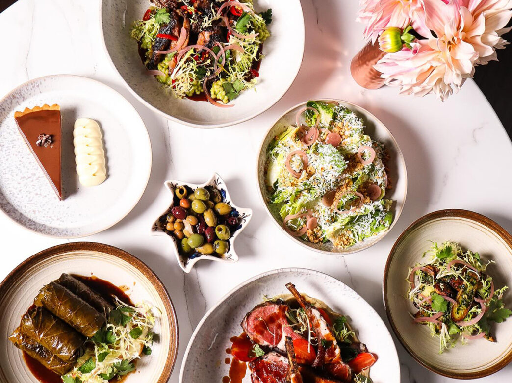 An overhead view shows a spread of plated dishes on a white table, including salads, stuffed grape leaves, a piece of pie and a small bowl of mixed olives.