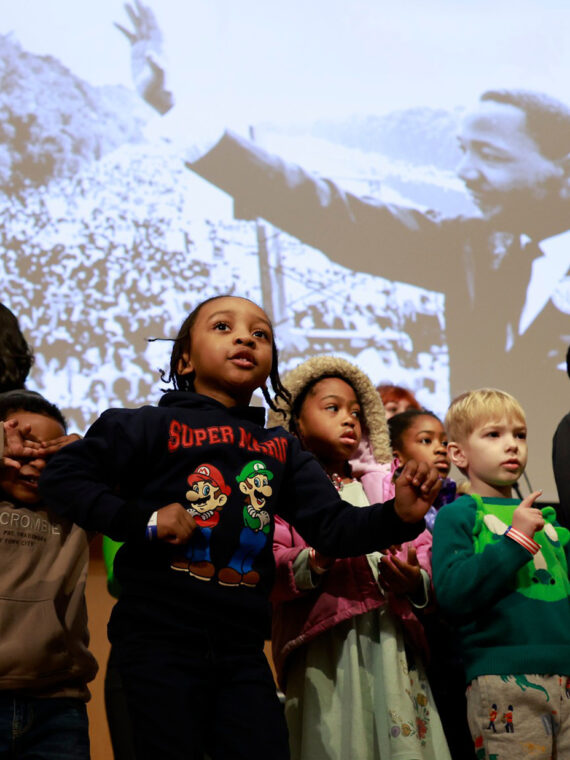 A group of children stands onstage in front of a large projected photo of Martin Luther King Jr., with several kids clapping and moving as part of a performance.