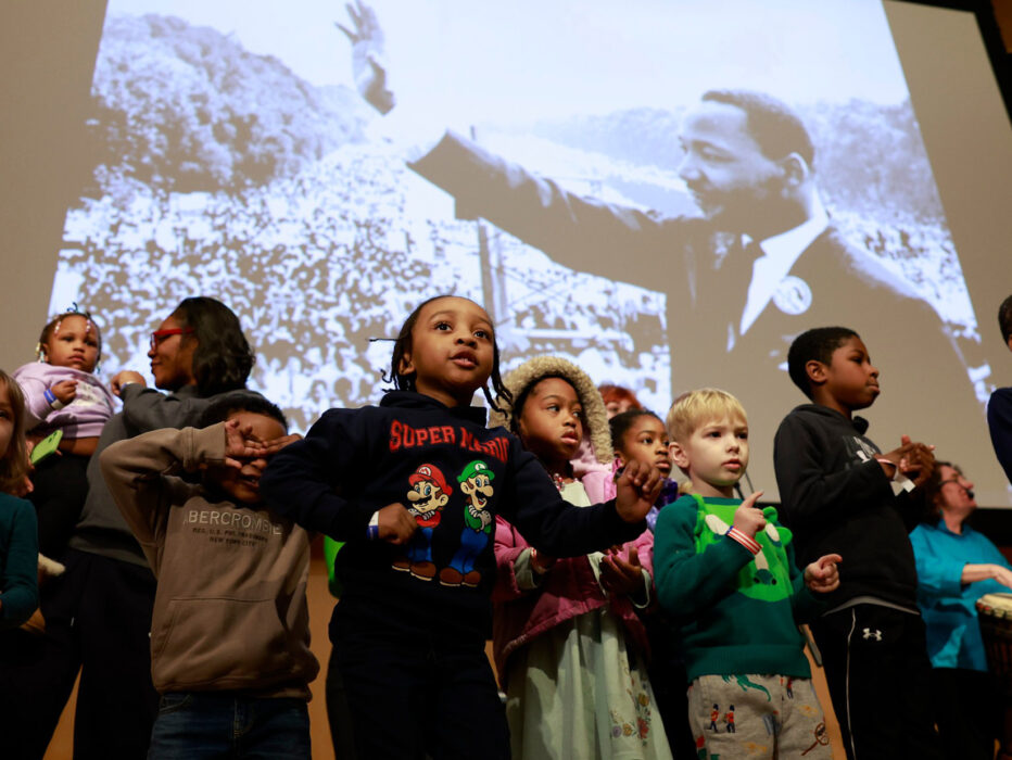 Um grupo de crianças fica no palco em frente a uma grande foto projetada de Martin Luther King Jr., com várias crianças batendo palmas e se movimentando como parte de uma apresentação.